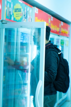 A person with a backpack is standing in front of a refrigerated display case in a store, possibly a convenience store or supermarket. Various products are visible inside the refrigerator, including bottles and packaged goods. The scene is lightly blurred with a cool tone.