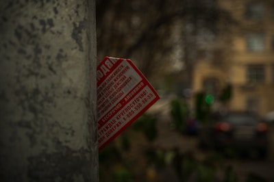 A weathered, gray concrete pole partially obscures a red and white flyer with text, set against an out-of-focus urban background featuring building facades and foliage.