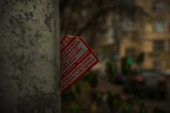 A weathered, gray concrete pole partially obscures a red and white flyer with text, set against an out-of-focus urban background featuring building facades and foliage.