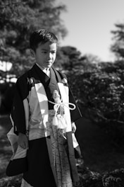 A young boy is standing outside in a formal, traditional garment with intricate patterns. The background features blurred foliage and trees, adding a natural element to the setting.