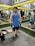 A person stands in a gym wearing a blue tank top, black shorts, and gloves. They are positioned next to a weightlifting belt on a bench. The gym has mirrors on the walls, exercise equipment in the background, and yellow accents.