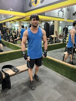 A person stands in a gym wearing a blue tank top, black shorts, and gloves. They are positioned next to a weightlifting belt on a bench. The gym has mirrors on the walls, exercise equipment in the background, and yellow accents.