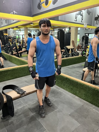 A person stands in a gym wearing a blue tank top, black shorts, and gloves. They are positioned next to a weightlifting belt on a bench. The gym has mirrors on the walls, exercise equipment in the background, and yellow accents.