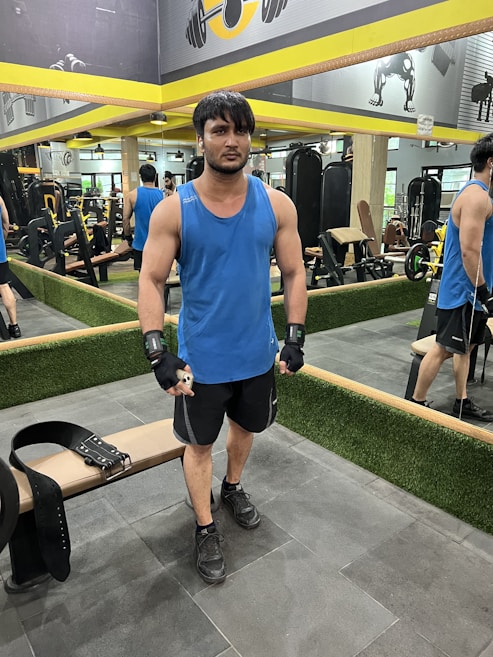 A person stands in a gym wearing a blue tank top, black shorts, and gloves. They are positioned next to a weightlifting belt on a bench. The gym has mirrors on the walls, exercise equipment in the background, and yellow accents.