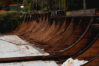 A series of wooden ramps in a skatepark, with a worn and weathered appearance. The area is surrounded by greenery, and there are some graffiti marks on the structure. The ramps and surrounding environment have an abandoned, untended look, giving a sense of decay over time.