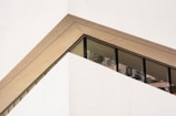 A modern architectural design featuring a diagonal section of a building with a prominent glass window. The structure has clean, sharp lines and a geometric shape, with a beige frame surrounding the window.