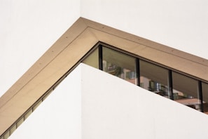 A modern architectural design featuring a diagonal section of a building with a prominent glass window. The structure has clean, sharp lines and a geometric shape, with a beige frame surrounding the window.