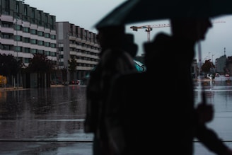 A rainy urban scene with a street reflecting the wet conditions. An outline of several people under umbrellas is visible in the foreground, while in the background there are apartment buildings and a construction crane. The street is largely empty except for a few cars and the overcast sky creates a dim, subdued light.