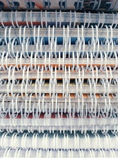 A close-up view of several spiral-bound notebooks stacked vertically, showcasing the overlapping rings and edges of the pages. The spirals are white, and various colors from the notebook covers and pages peek through the spaces between them.