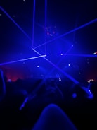 A dark room filled with people focused on a light show with blue lasers crossing the space. The atmosphere is vibrant and energetic, suggesting a concert or a club scene.