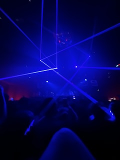 A dark room filled with people focused on a light show with blue lasers crossing the space. The atmosphere is vibrant and energetic, suggesting a concert or a club scene.