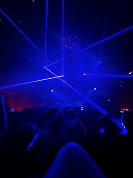 A dark room filled with people focused on a light show with blue lasers crossing the space. The atmosphere is vibrant and energetic, suggesting a concert or a club scene.