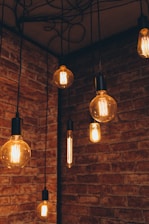 A series of vintage-style Edison bulbs hang from the ceiling against a backdrop of rustic red brick walls. The exposed filaments of the bulbs emit a warm glow, creating an inviting and cozy atmosphere.