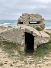 An old, weathered concrete bunker sits partially buried in the sand, overlooking the sea. The structure has an entrance at the front and a small, narrow window on top. The sky is overcast, and the surrounding landscape is covered with sparse grass and vegetation.