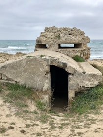 An old, weathered concrete bunker sits partially buried in the sand, overlooking the sea. The structure has an entrance at the front and a small, narrow window on top. The sky is overcast, and the surrounding landscape is covered with sparse grass and vegetation.