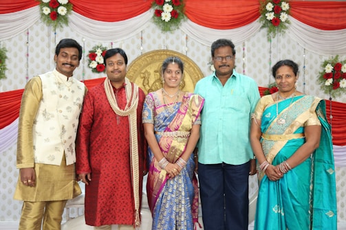 A group of five people dressed in colorful traditional Indian attire, standing in front of a decorated backdrop. The backdrop features red and white drapery with floral arrangements. A golden emblem is displayed at the center, surrounded by drapery and flowers.