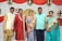 A group of five people dressed in colorful traditional Indian attire, standing in front of a decorated backdrop. The backdrop features red and white drapery with floral arrangements. A golden emblem is displayed at the center, surrounded by drapery and flowers.