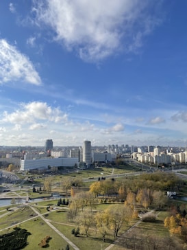 A sprawling urban landscape with a mix of modern high-rise buildings and open green spaces. The sky is a vibrant blue with scattered clouds, adding contrast to the city's skyline. The foreground features a well-maintained park with walking paths, trees, and a small body of water.