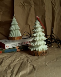 Two decorative candles shaped like Christmas trees are placed on a stack of books. The candles have bark-like bases and are surrounded by a string of fairy lights. Everything is set against a backdrop of crumpled kraft paper, giving the scene a cozy and rustic ambiance.