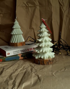 Two decorative candles shaped like Christmas trees are placed on a stack of books. The candles have bark-like bases and are surrounded by a string of fairy lights. Everything is set against a backdrop of crumpled kraft paper, giving the scene a cozy and rustic ambiance.