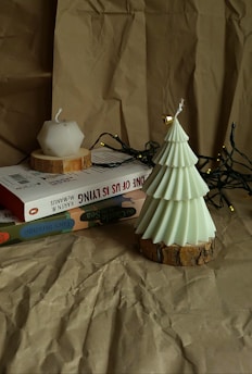 A geometric candle with a white hue is positioned on top of a stack of books, accompanied by a decorative candle shaped like a Christmas tree. The books have colorful covers, and a string of small fairy lights is casually draped around them. The background consists of crumpled brown paper, adding a textured and rustic feel to the scene.