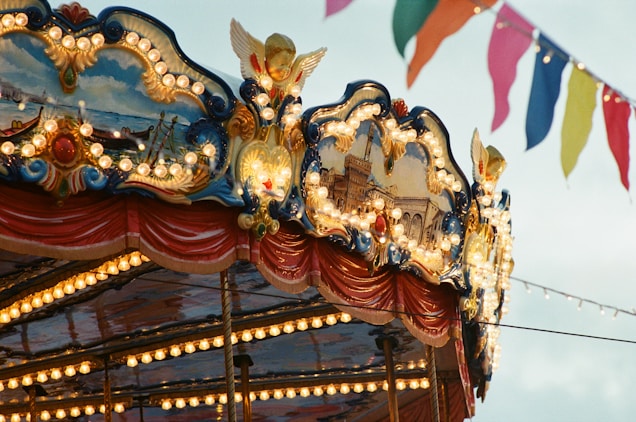 An ornate carousel top decorated with intricate paintings and glowing light bulbs. The design features classical architecture and maritime scenes. Colorful pennant flags flutter in the sky above, suggesting a festive atmosphere.