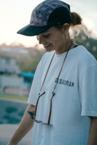 A person wearing a patterned baseball cap and a white t-shirt with text. A pair of sunglasses hangs from a strap around the neck. The individual appears to be smiling and is in an outdoor setting, possibly a park or open space, with a blurred background.