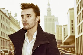 A person with styled hair and a serious expression is wearing a dark coat and a white shirt, walking in an urban environment. Tall buildings and a grand structure are visible in the background, indicating a cityscape with an architectural focus. The photo has a warm, vintage tone.