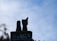 A bird is perched on a chimney against a backdrop of a cloudy sky. The chimney is made of brick, and there is a silhouette of trees visible at the bottom.