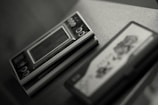 A close-up of a vintage handheld electronic game device sitting on a surface. The device features a small screen and several circular buttons, with partially visible branding. To the right, there is another object, possibly a game cartridge, with indistinct graphics.