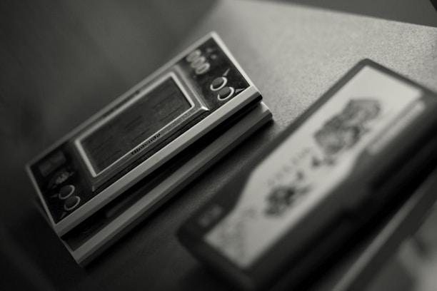A close-up of a vintage handheld electronic game device sitting on a surface. The device features a small screen and several circular buttons, with partially visible branding. To the right, there is another object, possibly a game cartridge, with indistinct graphics.
