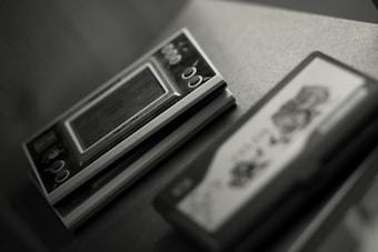 A close-up of a vintage handheld electronic game device sitting on a surface. The device features a small screen and several circular buttons, with partially visible branding. To the right, there is another object, possibly a game cartridge, with indistinct graphics.