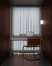 A compact workspace featuring a modern desk setup with a sleek laptop open to a design program. A simple chair with a metal frame is placed in front of the desk. The setting is minimalistic, enclosed by dark wooden walls, and softly illuminated by a wall-mounted lamp on the right. Floor-to-ceiling curtains cover a large window in the background, creating a cozy and private atmosphere.
