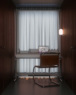 A compact workspace featuring a modern desk setup with a sleek laptop open to a design program. A simple chair with a metal frame is placed in front of the desk. The setting is minimalistic, enclosed by dark wooden walls, and softly illuminated by a wall-mounted lamp on the right. Floor-to-ceiling curtains cover a large window in the background, creating a cozy and private atmosphere.