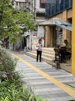 A city street scene with a person walking along a sidewalk lined with greenery. A man is seated using a sewing machine under an umbrella on a small platform next to a storefront. The area is urban with buildings, signs, and power lines visible.