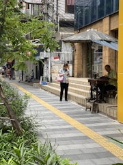 A city street scene with a person walking along a sidewalk lined with greenery. A man is seated using a sewing machine under an umbrella on a small platform next to a storefront. The area is urban with buildings, signs, and power lines visible.