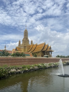 A large golden temple with intricate architectural details and a tall tower in the center is surrounded by well-manicured plants and a body of water with a small fountain. The sky is partly cloudy with patches of blue visible.
