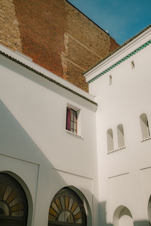 The image captures an architectural scene with white plaster walls featuring minimalist Moorish design elements like small, arched windows. A large brick wall provides a contrasting backdrop. The scene is bathed in natural light, casting sharp shadows on the walls. The sky is clear with a light blue hue.