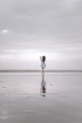 A solitary figure is seen in the distance on a vast, reflective beach, under an expansive, cloudy sky. The scene has a dreamlike and serene quality due to the out-of-focus element.
