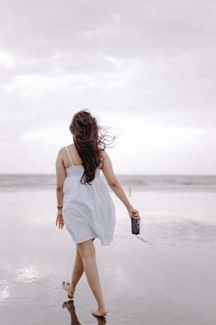 A candid shot of Hollan walking along a beach boardwalk, wearing a flowing white dress and simple leather sandals.