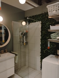 A modern bathroom with a glass shower enclosure and a rainfall showerhead. The room features a round mirror above a white sink with minimalistic beige toiletries. A stack of towels sits nearby on metal shelves which also hold a laundry basket. The walls are textured with green artificial plants.