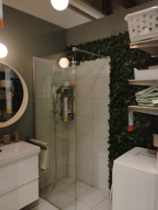 A modern bathroom with a glass shower enclosure and a rainfall showerhead. The room features a round mirror above a white sink with minimalistic beige toiletries. A stack of towels sits nearby on metal shelves which also hold a laundry basket. The walls are textured with green artificial plants.