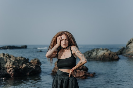 A woman with curly hair and tattoos stands on a rocky seashore. She is wearing a black crop top and skirt, with one hand on her hip and the other resting on her head. The sea is calm, and the sky is clear.