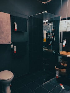 A modern bathroom showroom featuring a toilet, a glass shower enclosure, and dark tile flooring. The walls incorporate built-in cabinetry with minimalist designs. A wicker basket and some packaged products are visible under a counter.