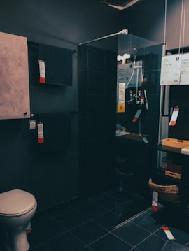 A modern bathroom showroom featuring a toilet, a glass shower enclosure, and dark tile flooring. The walls incorporate built-in cabinetry with minimalist designs. A wicker basket and some packaged products are visible under a counter.