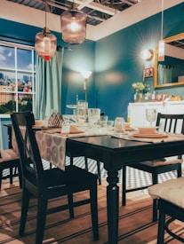 A contemporary dining room with a neatly set table is adorned with modern decor. The space features a teal color theme with dark wooden chairs and a table covered with a patterned runner. Hanging pendant lights and atmospheric table lamps illuminate the room, creating a cozy ambiance. A cityscape view is visible through the window, complementing the interior's stylish aesthetic. Decorative elements like a vase with flowers and framed pictures enhance the homely feel.
