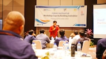 A group of people are attending a workshop focused on capacity building for instructors. The event is held in Padang and organized by various organizations, as indicated by the logos on the banner. A speaker in a red shirt is addressing the audience from a podium, while participants sit at round tables with water bottles and notes in front of them.