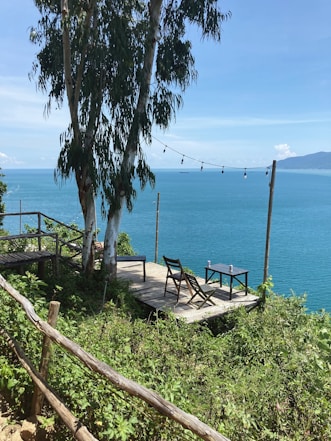 A serene wooden deck is situated on a hillside overlooking a vast, calm blue ocean. The deck is equipped with two wooden chairs and a small table, with hanging lights strung between wooden posts. Lush green foliage surrounds the area, and tall eucalyptus trees provide shade. The background includes a view of distant mountains under a clear blue sky.