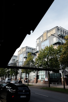 A modern multi-story office building with large windows and metal framing is visible. Trees line the sidewalk in front of the building. A pedestrian bridge connects different parts of the building above the street. Several vehicles are parked along the road, and a few people are walking on the sidewalk.