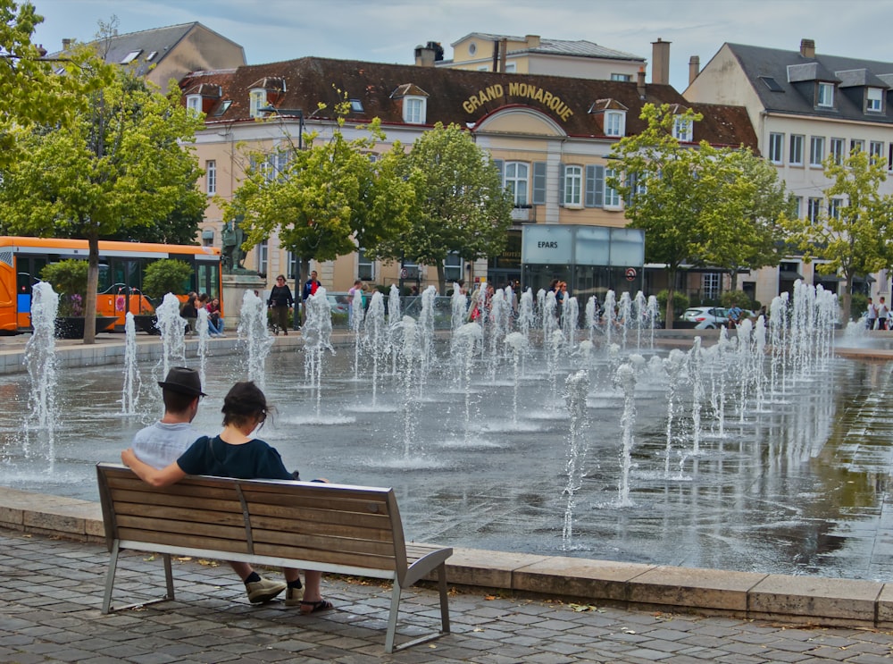 two people sitting on a bench in front of a fountain